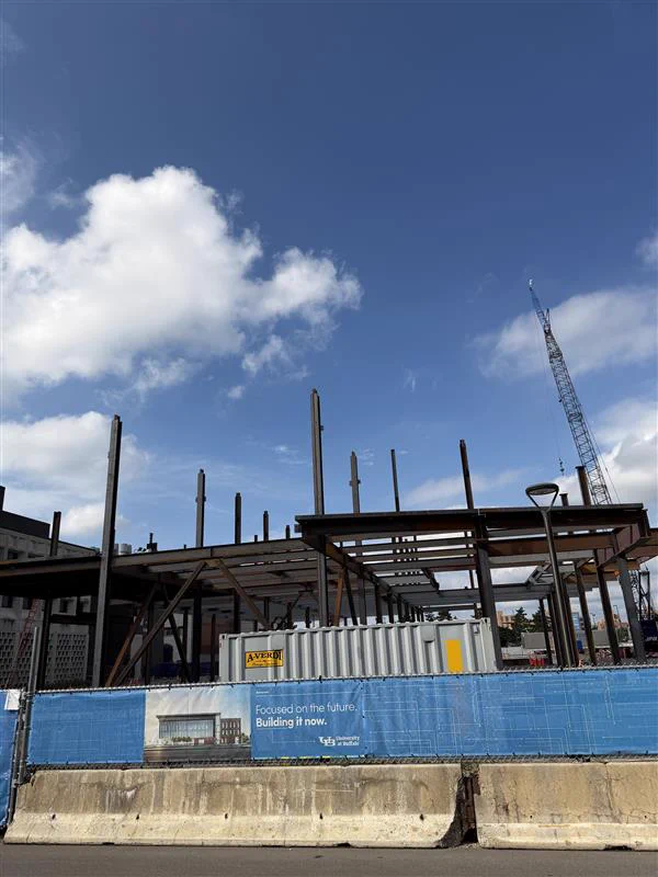 Steel skeleton of a new building under construction against a bright blue sky with white clouds. A large crane is visible in the background. In the foreground, a concrete barrier and a blue construction fence display text: \"Focused on the future. Building it now. University at Buffalo\"