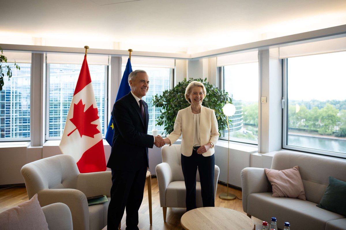 Prime Minister Mark Carney and the President of the European Commission, Ursula von der Leyen, shake hands while smiling for a photo ahead of their meeting in Strasbourg, France. The flags of Canada and the European Union are behind them.