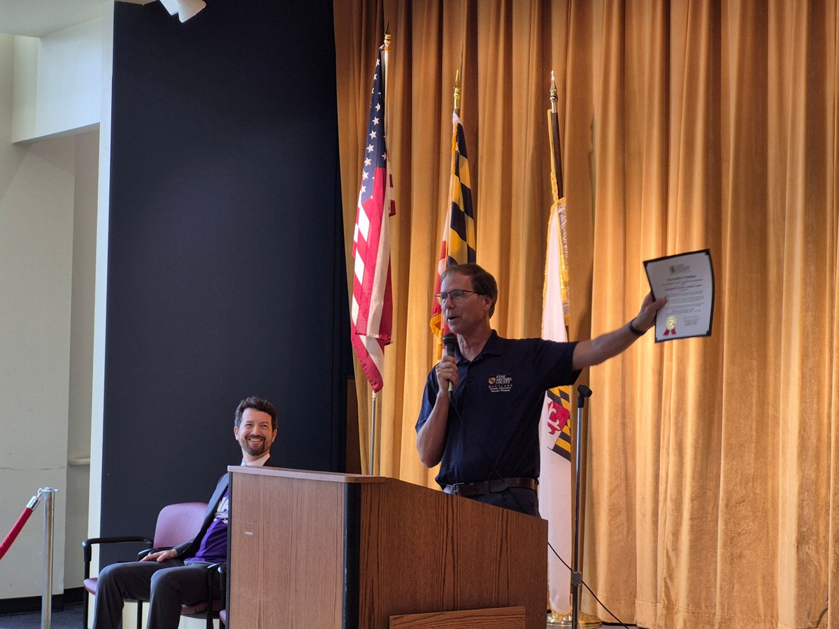 County Executive Pittman stands at a podium holds up a citation and speaks into a microphone while Mayor Littmann sits beside him, with U.S. and state flags and gold stage curtains behind them.