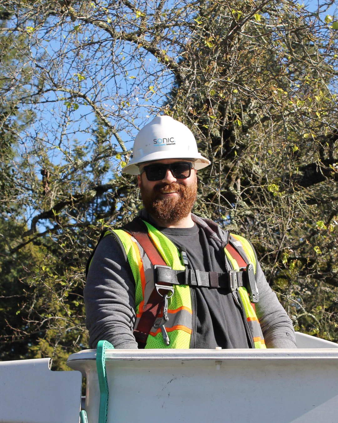 Casey, a Sonic Fiber Internet Drop Lead, wearing a hard hat, safety vest, and fall-protection harness, stands in an aerial bucket lift outdoors near tree branches. 