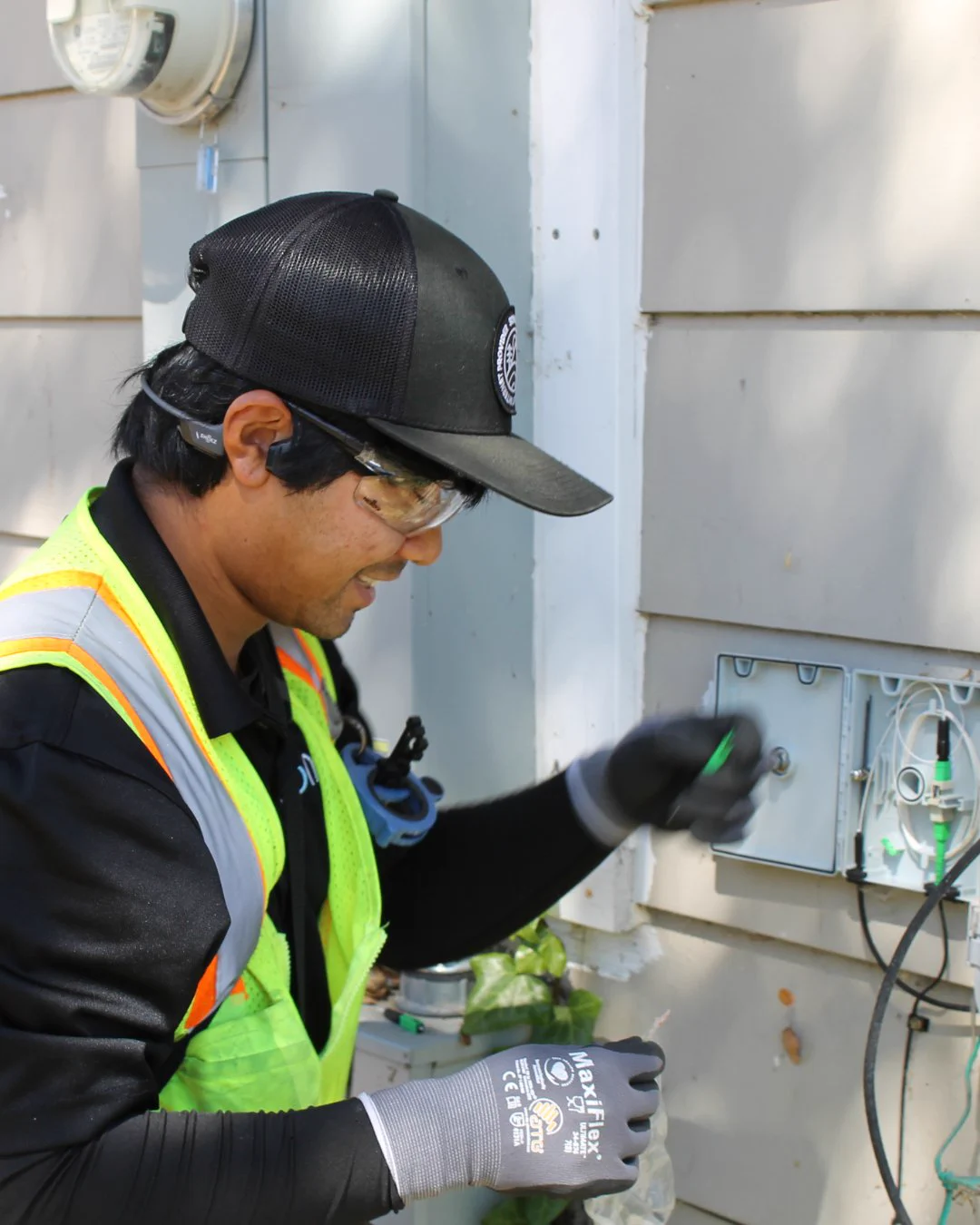 Randy, a Sonic Fiber Internet Technician, wearing a high-visibility safety vest, safety glasses, gloves, and a Sonic Fiber Internet-branded hat, is smiling and working with the fiber line and a keystone outdoors. The transition box that's mounted on the outside of a home is open as Randy works. 