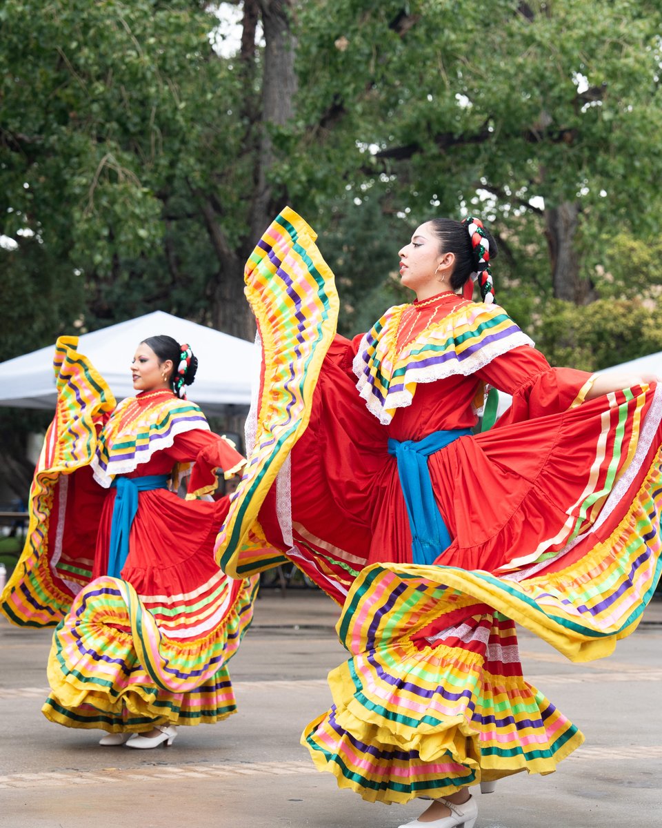 Two people performing a traditional folk dance, wearing vibrant red dresses with colorful ruffles and blue sashes. Their outfits include decorative hairpieces as they gracefully move, with green trees and white canopies in the background.