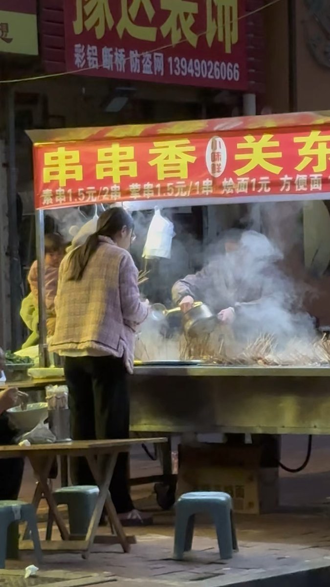ASchrimpf's tweet photo. A random mid-shot of a street food vendor flipping grilled skewers at a dusk roadside stall, soft warm neon light spilling over the pavement, passersby lingering for a quick bite. https://t.co/Pcr6UZ3PZJ