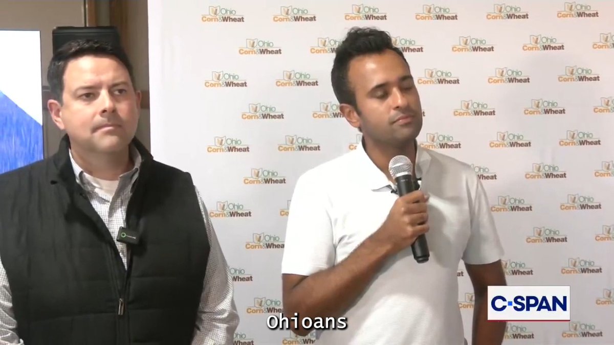 Two men stand before an "Ohio Corn & Wheat" backdrop during what appears to be a press event, with one man holding a microphone while speaking, as C-SPAN displays the closed caption "Ohioans." The scene corresponds to a reporter's question about Republican criticism regarding private jet use, highlighting the political discussion around candidate Vivek Ramaswamy and affordability concerns in Ohio.