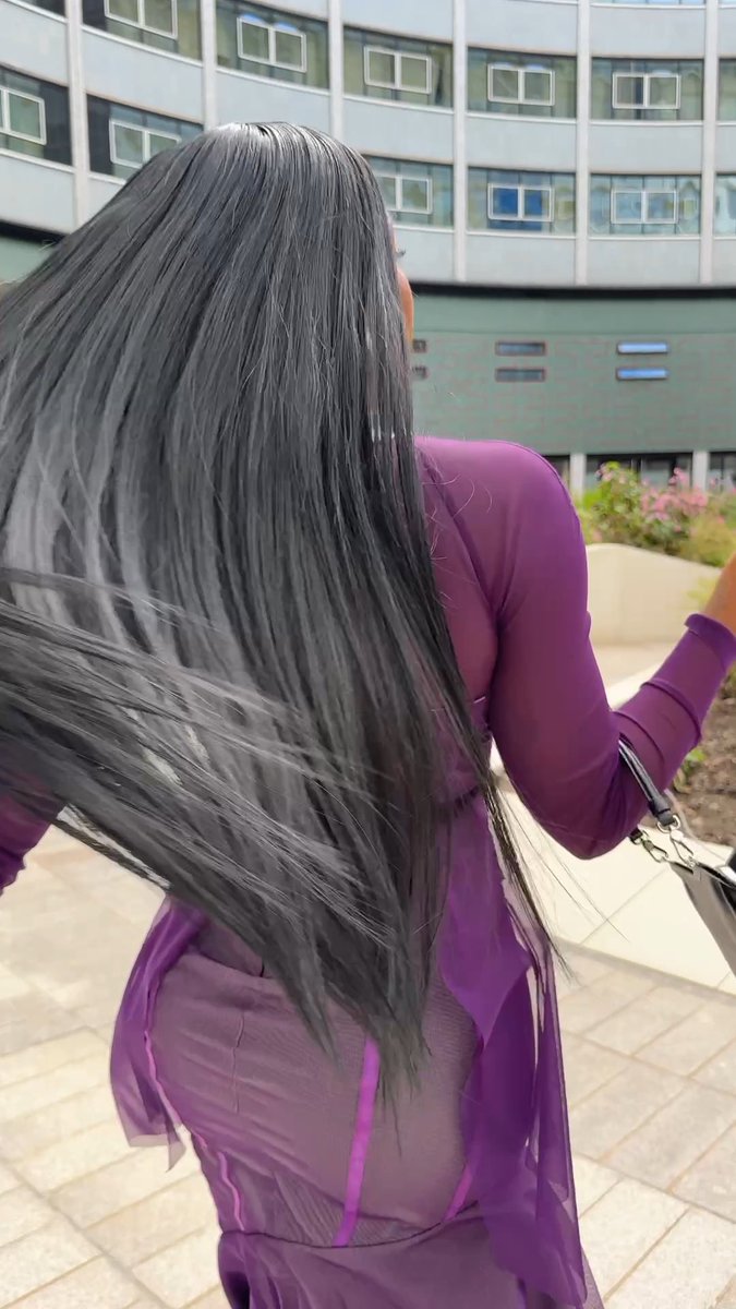 A woman with long, flowing gray hair stands on a balcony overlooking a city street, wearing a purple mesh dress.