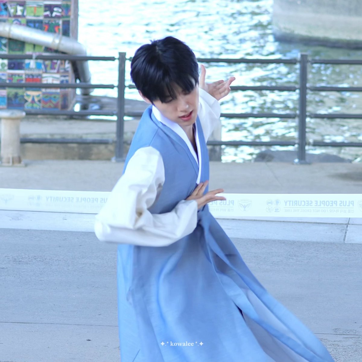 A young man in a light blue and white traditional Korean hanbok performing a dance pose outdoors near a waterfront, with the watermark 'kowalee' visible.