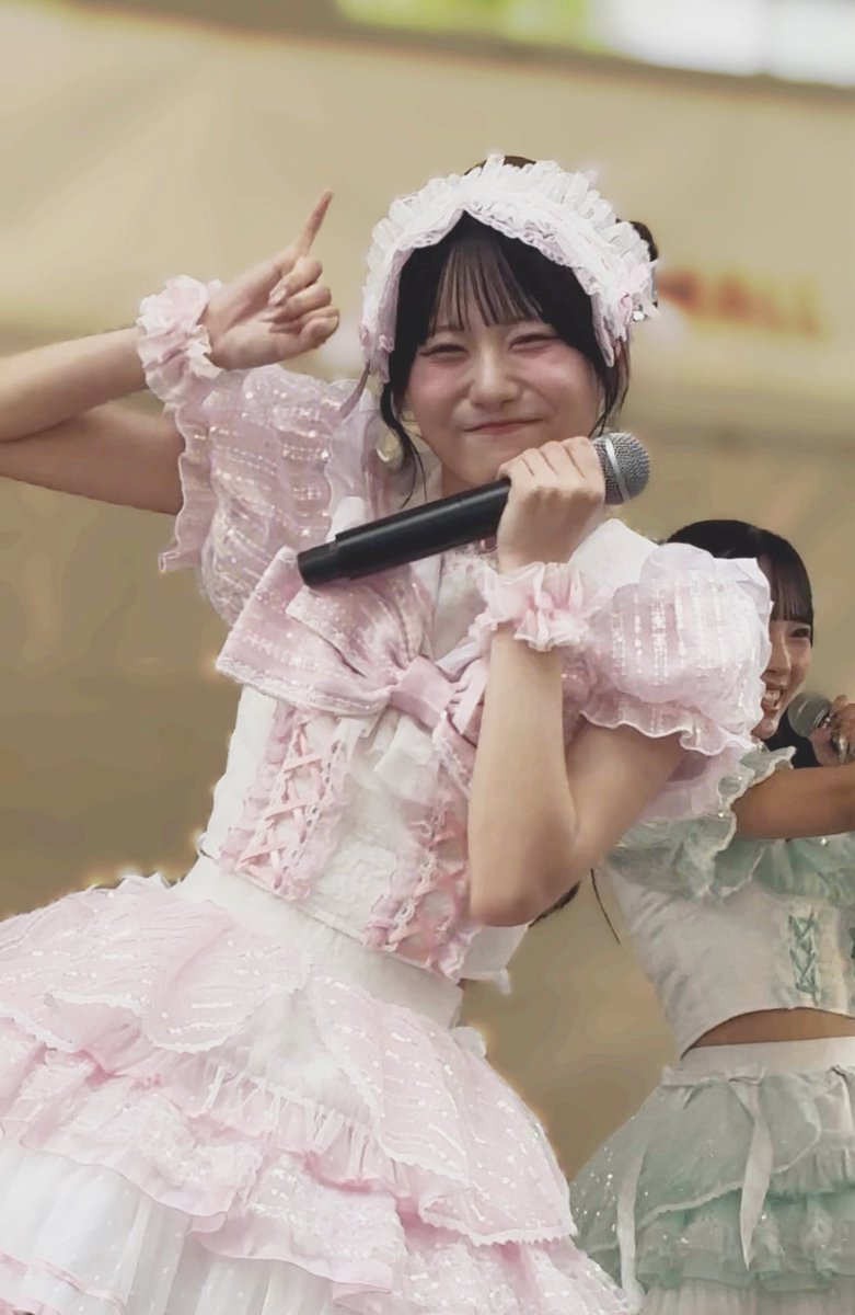 A young woman in a pink and white frilly maid-style outfit with a lace headband smiles and winks while holding a microphone, performing on stage with another member visible in the background wearing green.