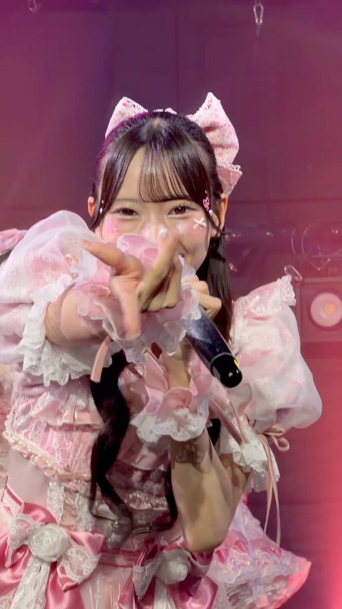 A Japanese pop idol performs on stage wearing an elaborate pink frilly dress with lace details, a matching hair bow, and decorative face stickers, pointing toward the camera while holding a microphone.
