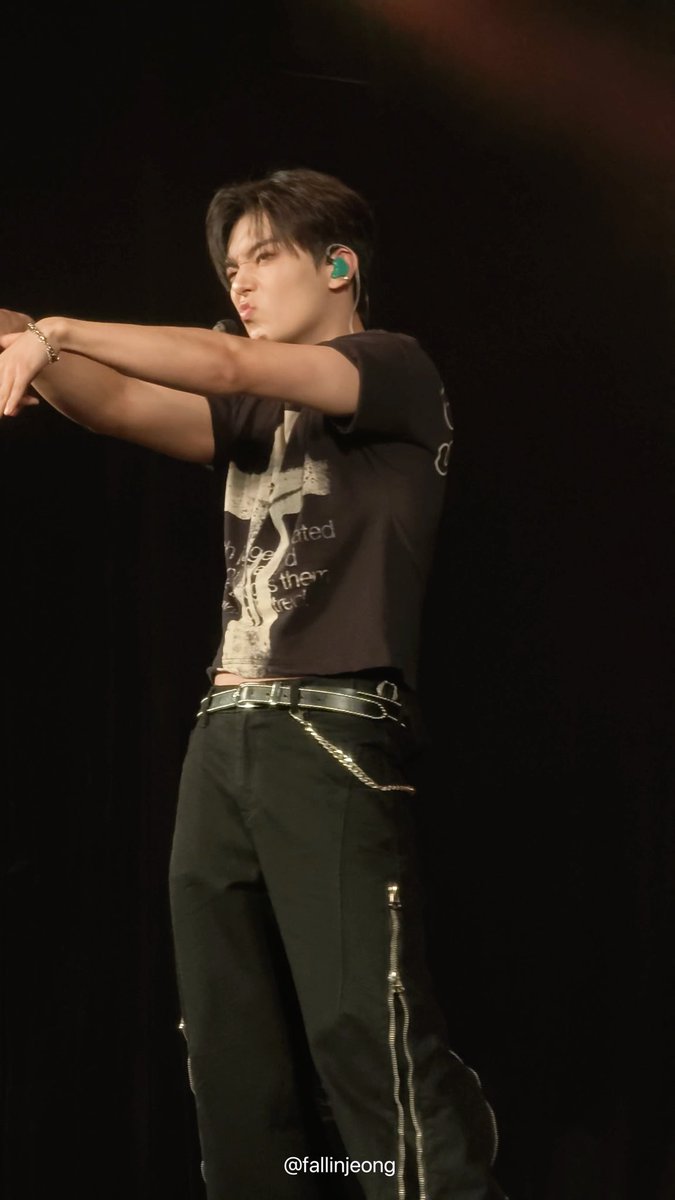 A male performer on stage wearing a black graphic t-shirt and black pants with zippers and a chain, extending his arms forward with a green in-ear monitor visible. Watermark reads @fallinjeong.
