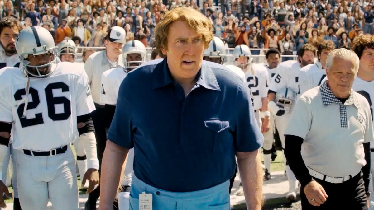 A scene from a football film showing a coach walking onto a field flanked by players in white and silver uniforms and an older man in a polo shirt.