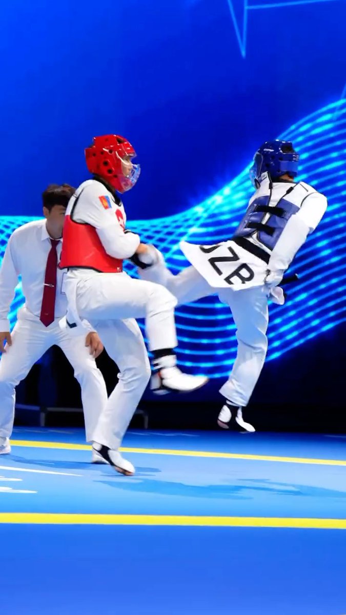A taekwondo sparring match in progress on a blue mat, with a competitor in red headgear and chest protector facing off against an opponent in blue headgear performing a high kick, with a referee in white shirt and red tie observing.