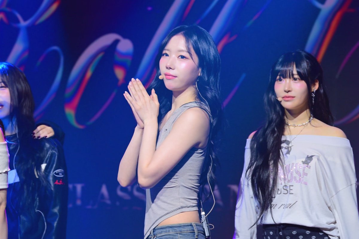 Three women perform on a stage lit in blue, with the center woman wearing a grey tank top and holding her hands together in a praying gesture.