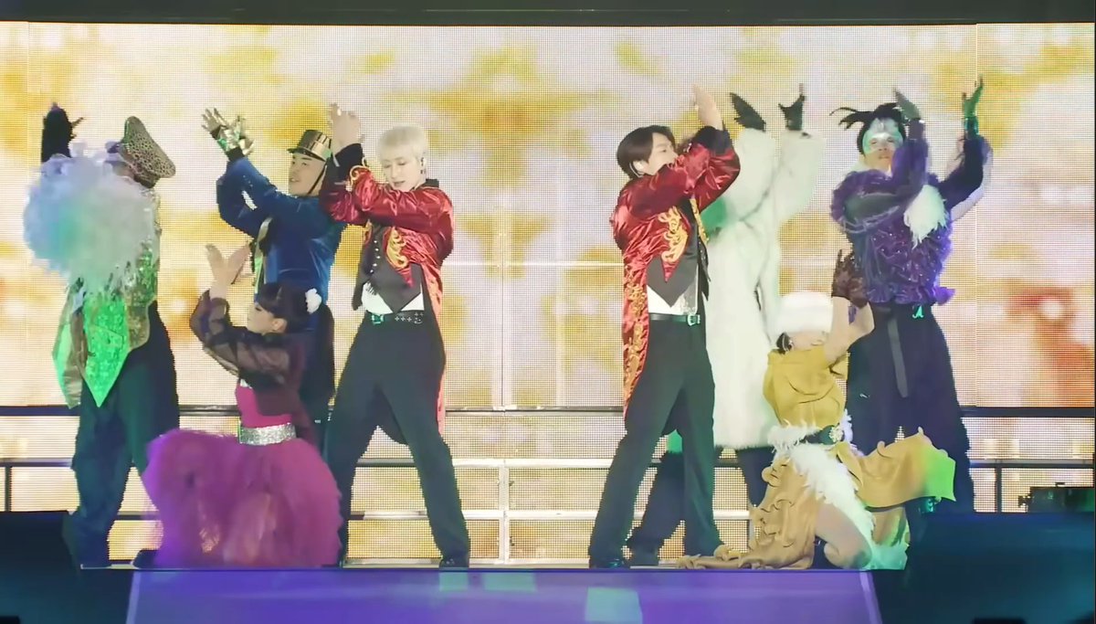 A group of performers in elaborate, colorful costumes on stage, featuring ringmaster-style red jackets and various theatrical outfits, performing what appears to be a K-pop or variety show performance with a yellow background screen.