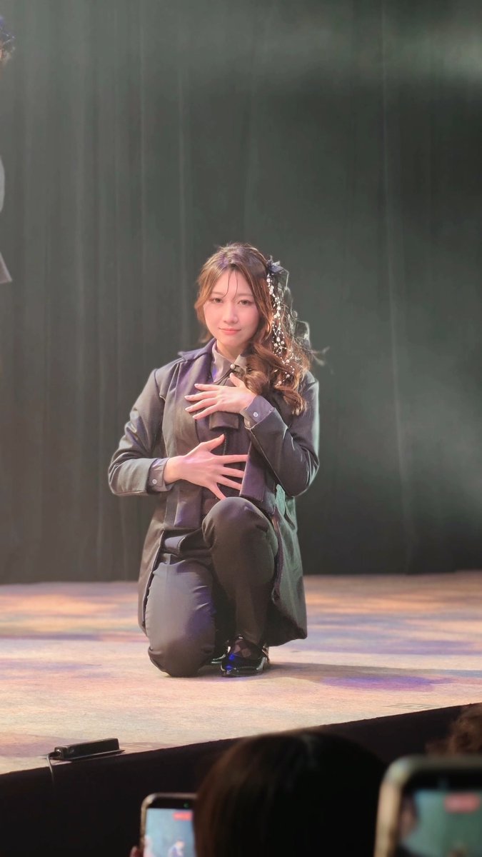 A young woman in a gray trench coat kneels on a stage with her hand over her heart, looking toward the audience under soft theatrical lighting.