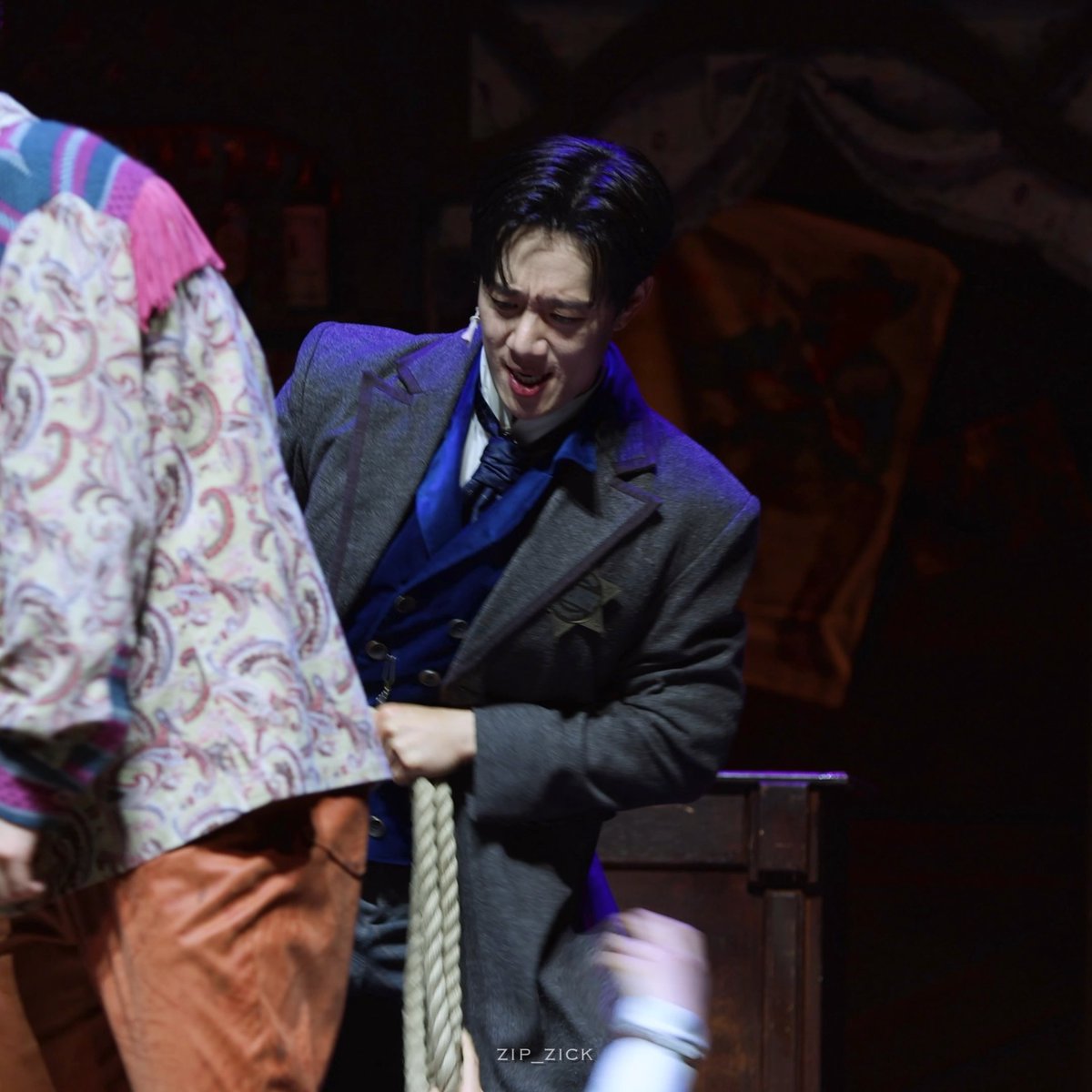 A theatrical performance photo showing a man in a grey coat with a blue sash and a Star of David pin laughing while holding a rope, with the watermark "ZIP_ZICK" visible.
