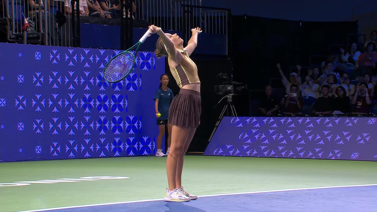 A female tennis player in a beige crop top and brown pleated skirt stands on a blue court, preparing to serve with her racket raised, while a ball girl and spectators watch in the background.