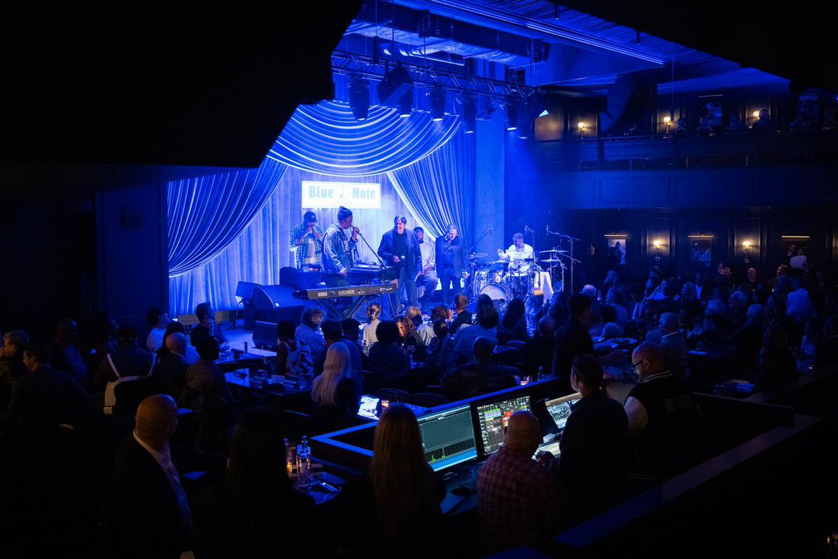 The interior of Blue Note London during its opening, with musicians performing on a brightly lit stage beneath blue lighting and draped curtains. The Blue Note sign is visible above the stage, while an audience fills the seating area in front.
