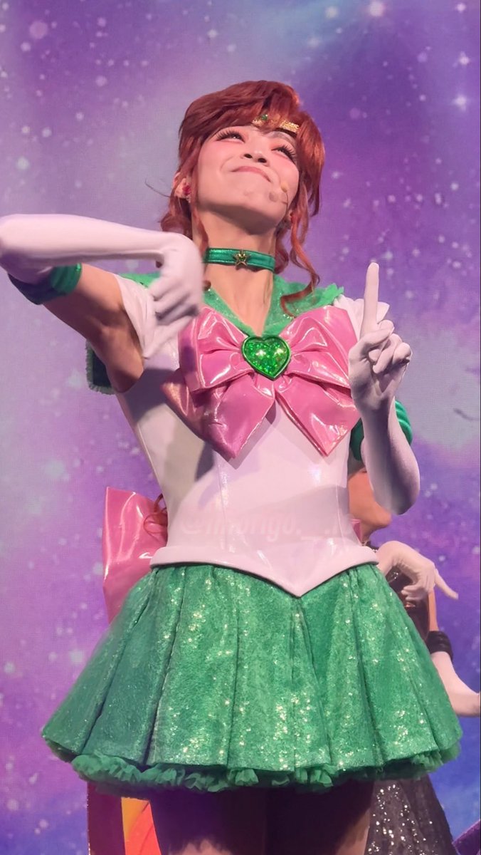 A performer dressed as Sailor Jupiter from Sailor Moon in her signature green and white costume with a large pink bow and white gloves, posing on a stage with a starry purple background.