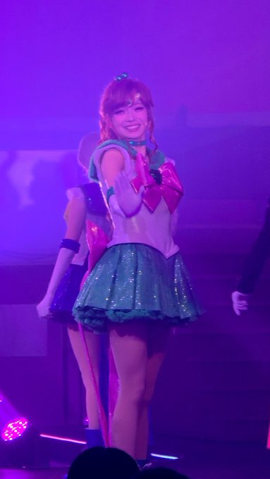 A performer on stage wearing a Sailor Moon-inspired costume with a white top, pink heart accents, and a sparkly blue skirt, posing with her arms raised under purple stage lighting.