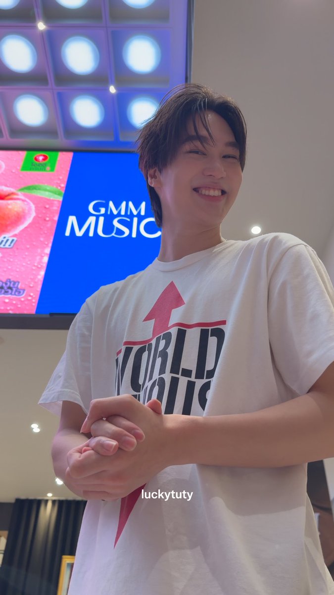 A smiling young man wearing a white 'World Tour' t-shirt stands in front of a screen displaying 'GMM MUSIC' and a peach beverage ad.