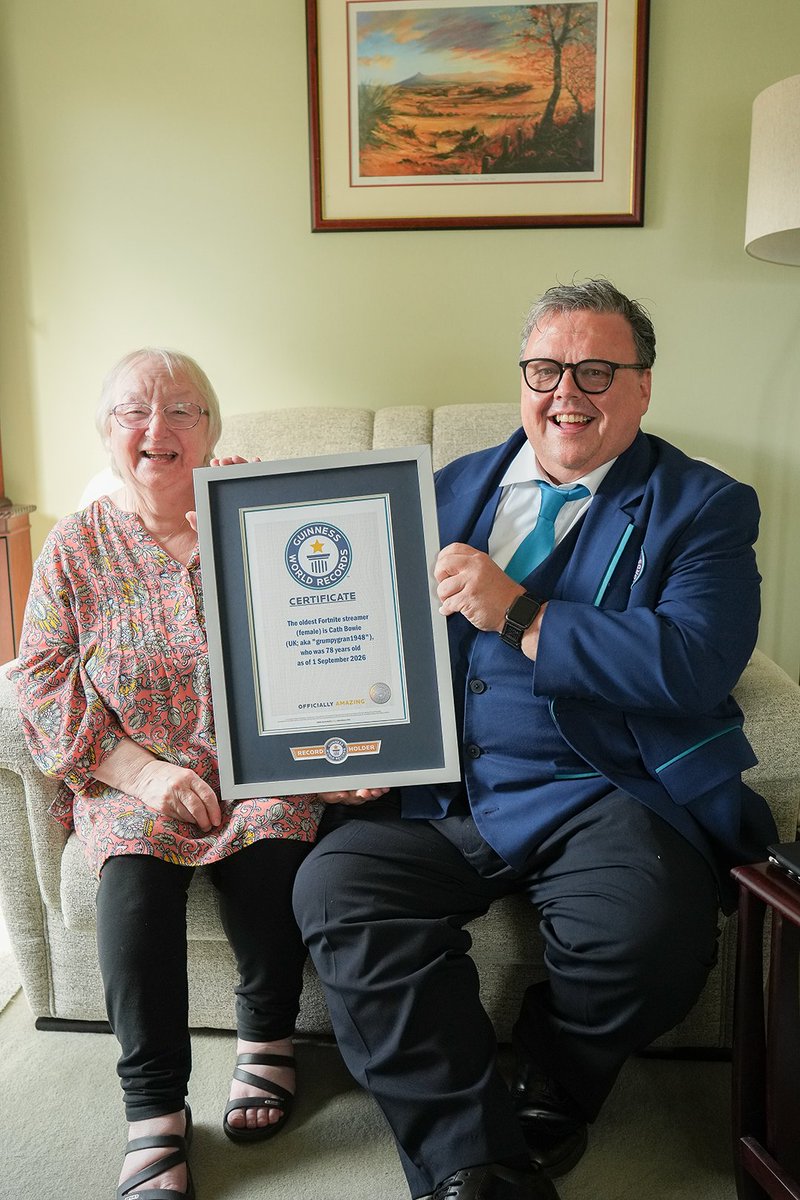 A woman and man sit on a couch smiling while proudly holding a framed Guinness World Records certificate. The certificate reads "The oldest Fortnite streamer (female) is Cath Bowie (UK, aka 'grumpygran1948'), who was 78 years old as of 1 September 2026," celebrating her recognition in the #GWRgamers 2027 book.