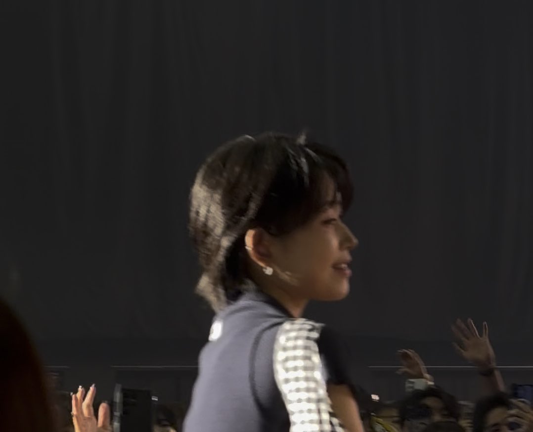 A young woman with short dark hair is shown in profile, wearing a checkered-patterned top with what appears to be an earpiece or headset microphone, set against a dark stage background with hands of an audience visible in the foreground. The Japanese caption expresses the poster's admiration for her beautiful side profile, referring to her as "Momona" (likely the idol Momoka Ito from the Japanese idol group = LOVE), while wondering if someone they reacted to recognizing might be a friend or just a fellow fan.