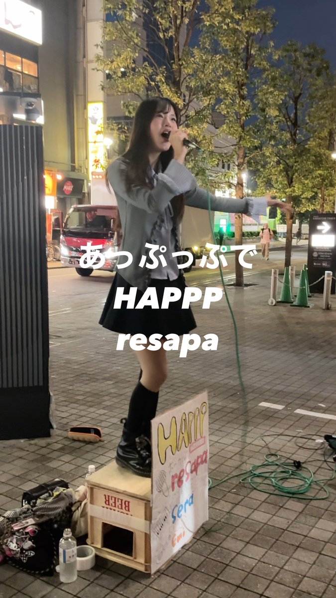 A street performer stands on a wooden crate singing into a microphone at night, wearing a gray ruffled cardigan and black skirt, with Japanese text 'あっぷっぷで' and 'HAPP resapa' overlaid.
