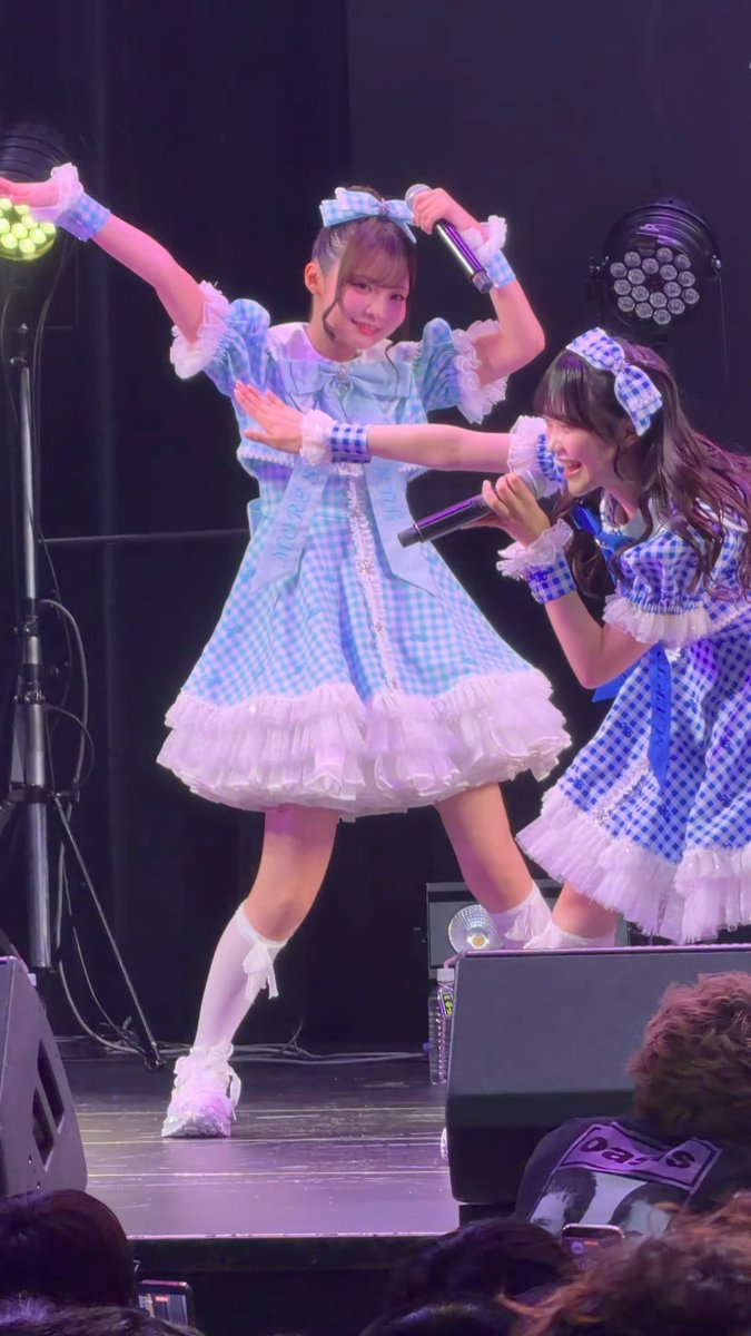 Two performers on stage wearing matching blue and white gingham-patterned idol outfits with frilly skirts, bows, and white knee-high socks. They are performing energetically, holding microphones, with stage lighting visible in the background.