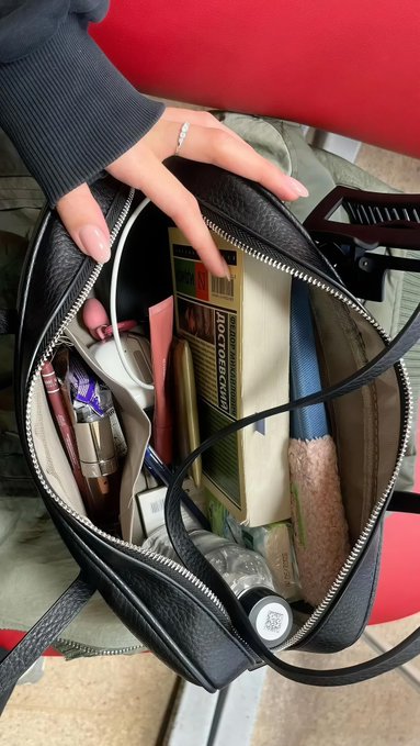 A hand reaches into an open black leather bag filled with books, cosmetics, and other personal items, including what appears to be a book titled 'AVICCENZA'.