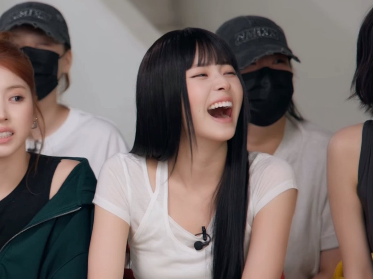A candid photo of a young woman with long black hair laughing joyfully while seated next to others wearing masks and caps.