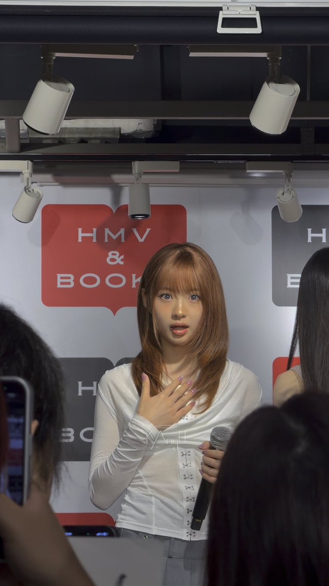 A young woman with reddish-brown hair speaks into a microphone at an HMV & Books event, wearing a white long-sleeved top.
