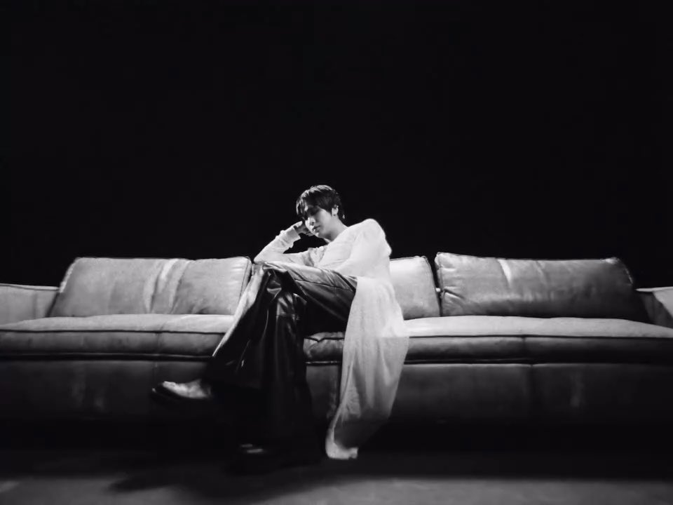 A man with dark hair sits contemplatively on a large leather couch, wearing a flowing white shirt over dark trousers, in a moody black-and-white setting with dramatic lighting. The image serves as a promotional artist visual for HAECHAN's first album "TASTE," with the caption "260924 artist visual SM instagram update with #HAECHAN #HAECHAN_TASTE."