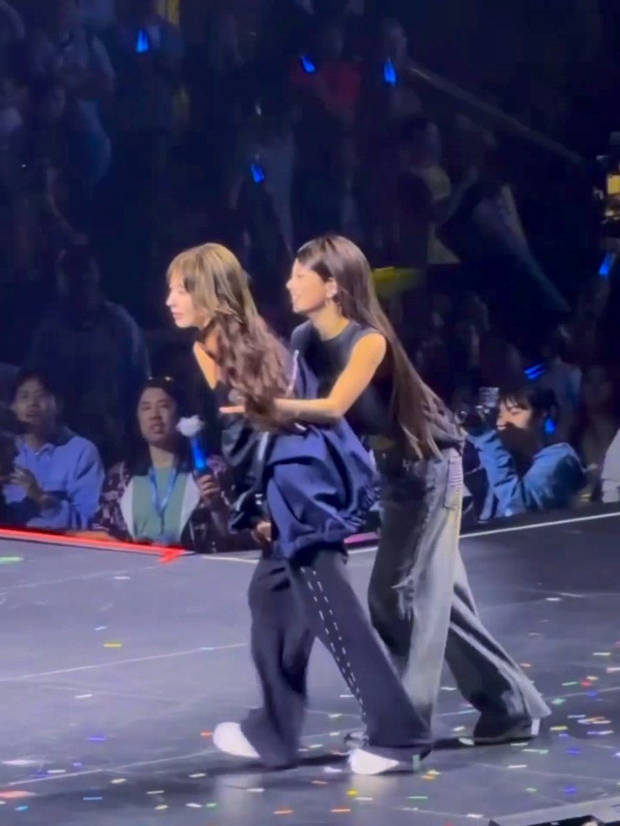 Two female performers walk across a stage scattered with confetti, with one playfully holding the other from behind, as fans in the background hold glowing blue light sticks. The intimate, joyful moment between the two performers embodies the sweet sentiment expressed in the caption.