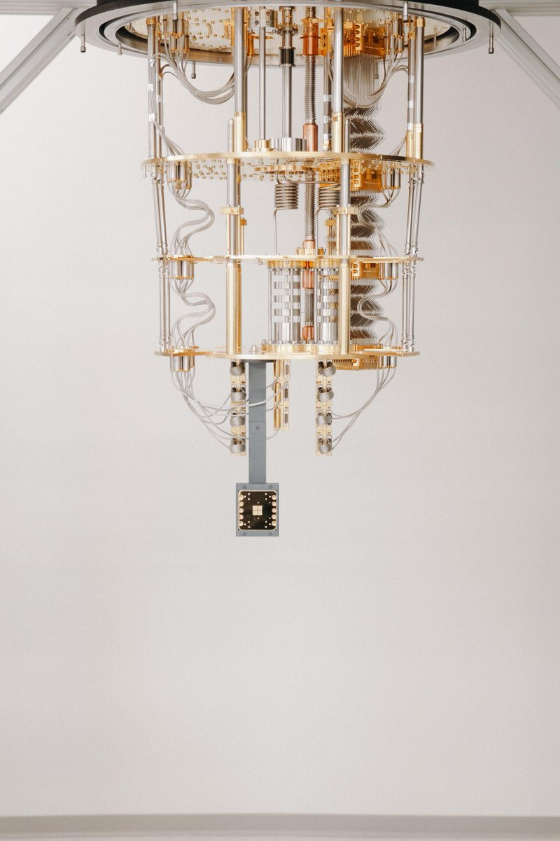 Close-up of a gold and silver quantum computing assembly hanging from the ceiling. It has stacked round plates, coiled tubes and bundles of wiring, with a small chip package at the bottom.