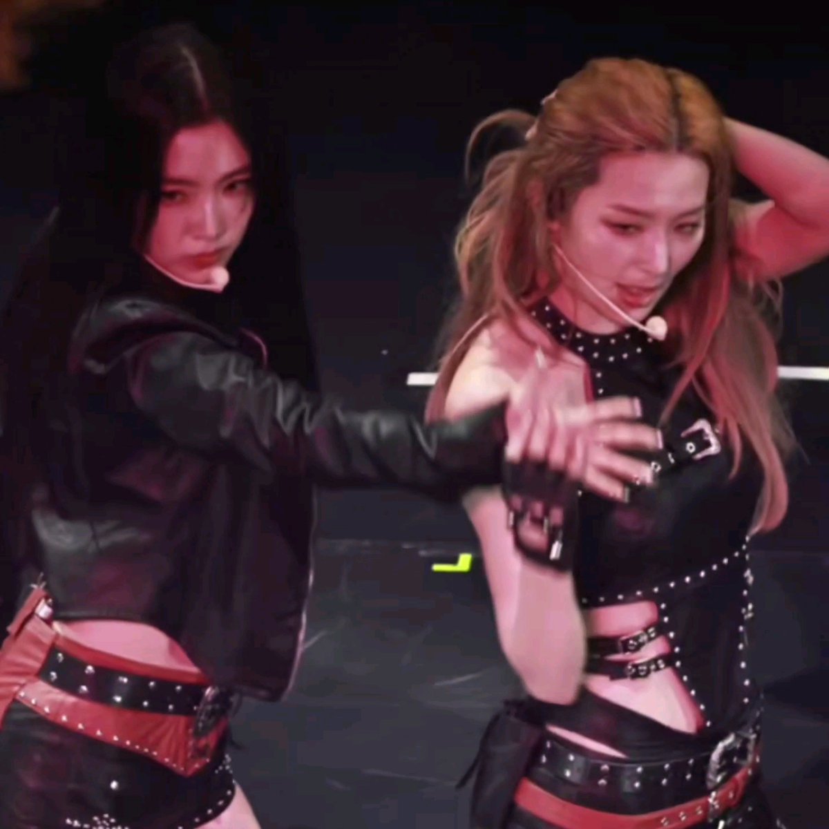 Two female K-pop performers on stage, both wearing black and red studded leather outfits with headset microphones.