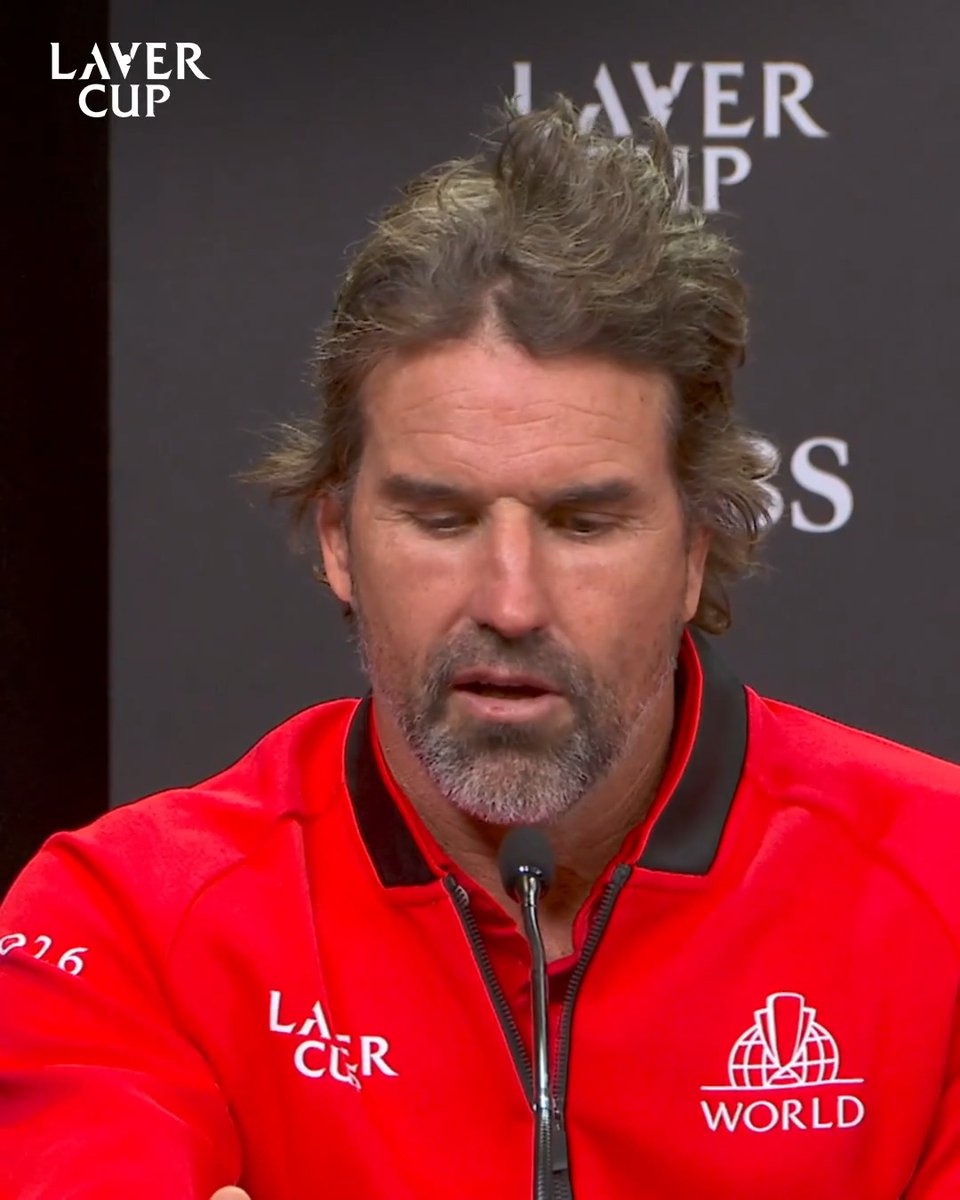 A close-up of a man with grayish-brown hair and a beard wearing a red Laver Cup jacket, speaking at a press conference, with the 'LAVER CUP' logo visible in the background.