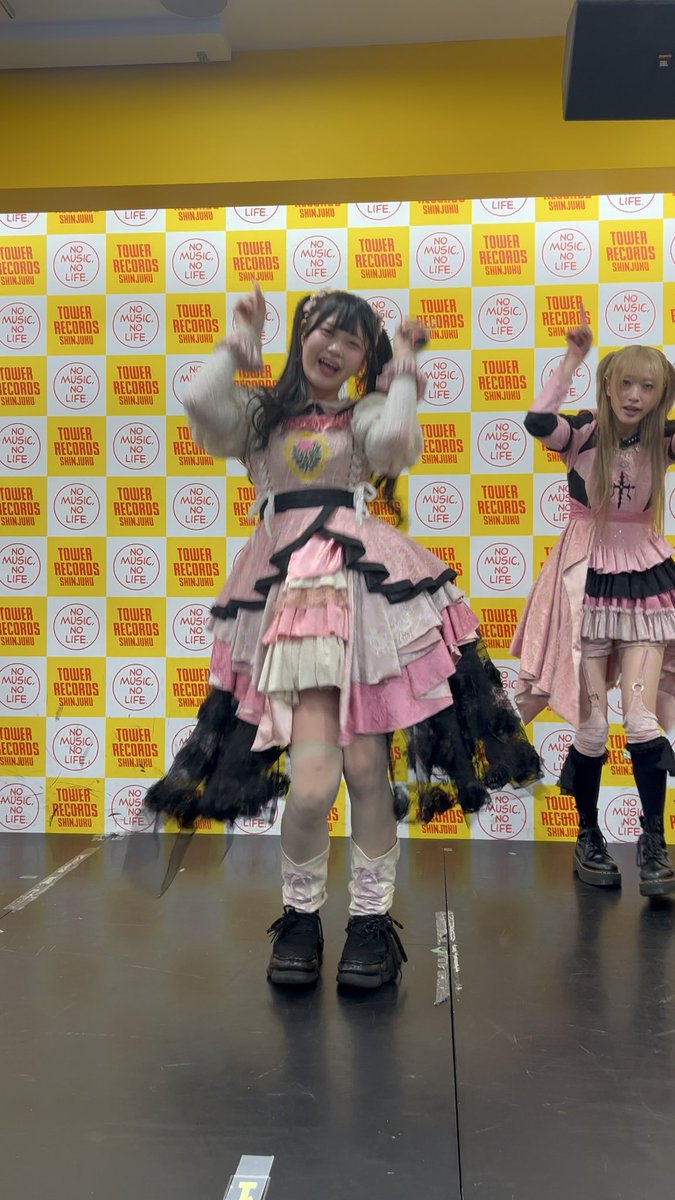 Two performers in pink and black frilly outfits pose enthusiastically on stage in front of a Tower Records 'NO MUSIC NO LIFE' backdrop featuring 'SHINJUKU' branding.