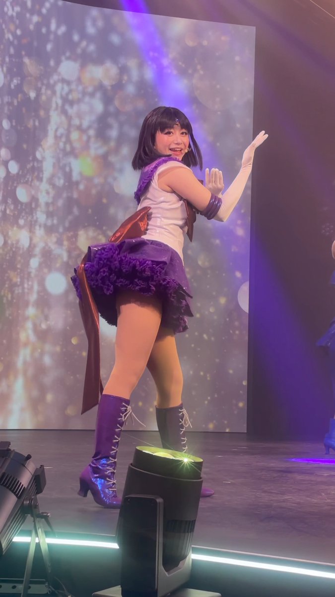 A performer on stage in a Sailor Moon-inspired purple and white costume with matching knee-high lace-up boots, posing in front of a sparkly LED backdrop.