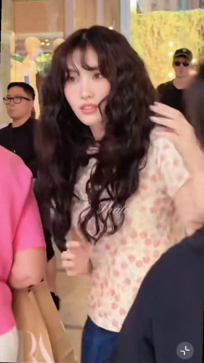 A young woman with long wavy dark hair and bangs wearing a floral top in what appears to be a mall or public indoor setting, with a watermark visible on the image.