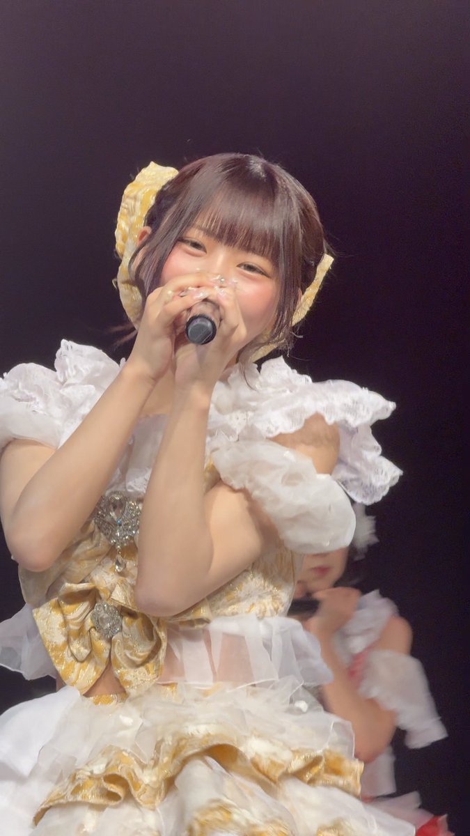 A close-up photo of a Japanese idol (resembling a member of a major idol group) performing on stage in an ornate white and gold dress while singing into a microphone.