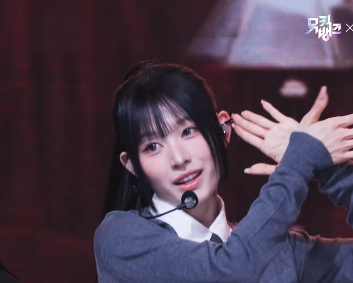 A young woman with long black hair and bangs, wearing a gray sweater over a white collared shirt and a headset microphone, smiles brightly while holding her hands up near her face in a playful gesture. In the top right corner, Korean text reads "뮤뱅" (Music Bank), indicating this is a screenshot from a K-pop music show performance. The caption references "Runaway" and "Minami's intro," suggesting this moment captures the energetic opening of Minami's "Runaway" stage appearance.