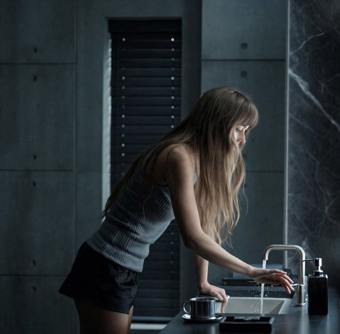 A woman with long hair stands at a sink in a dark, modern bathroom, washing her hands under a running faucet.