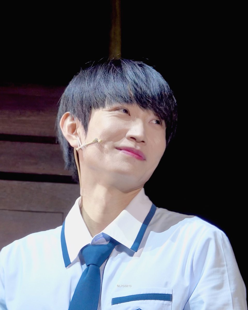 A young man with dark hair wearing a white shirt with blue trim and a blue tie smiles on stage, sporting a headset microphone.