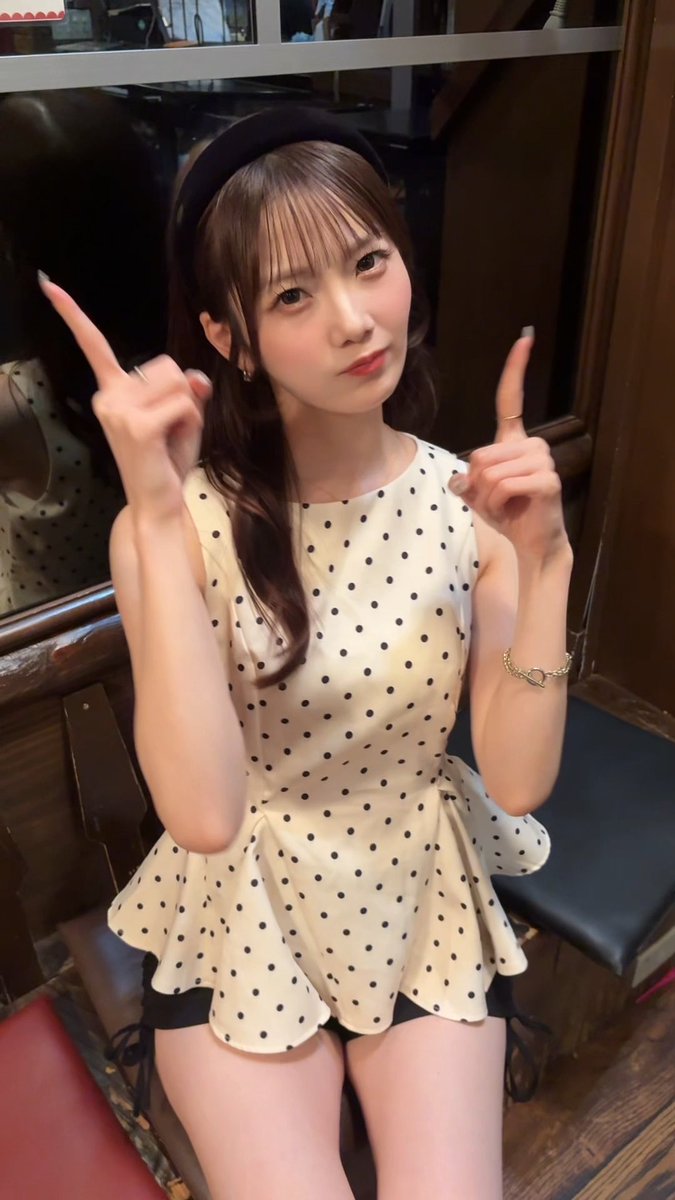 A young woman with a black headband and a white polka dot peplum top posing and pointing upwards with both index fingers.