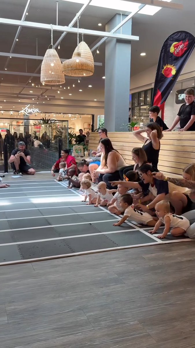 A baby crawling race event is taking place in a mall, with a line of infants and their parents waiting on a matted track while a photographer captures the scene.