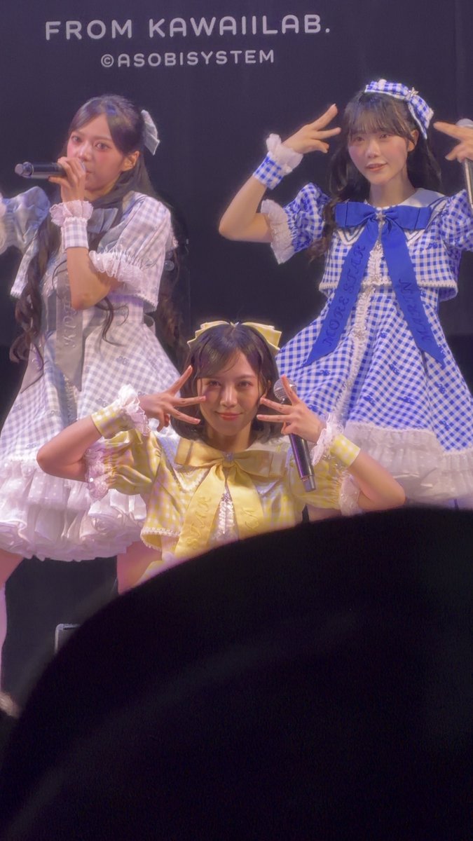 Three performers in blue and yellow gingham outfits perform on stage, with 'FROM KAWAIILAB.' and '©ASOBISYSTEM' displayed on the screen behind them.