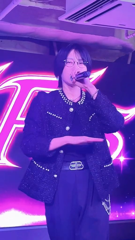 A performer on stage sings into a microphone, wearing a black tweed jacket with pearl detailing, a black top, and black pants, set against a purple-lit background with a pink logo.