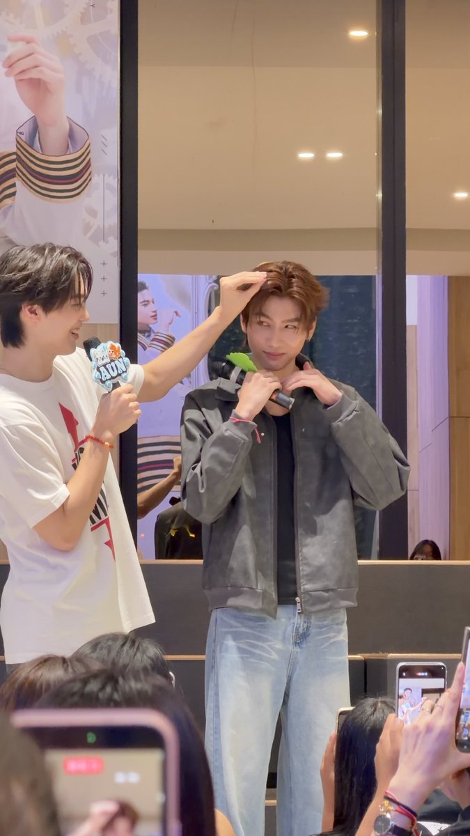 Two young men at what appears to be a fan event, one wearing a white t-shirt holding a microphone with a 'SUNNY' sticker, while the other in a gray jacket holds a toy microphone as fans take photos with phones.