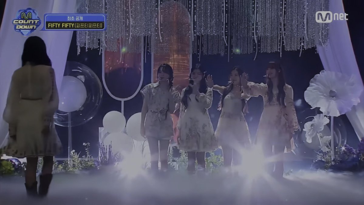 A screenshot from Mnet's M Countdown shows a K-pop group performing 'FIFTY FIFTY (피프티피프티)' on a stage with white decorations and fog.