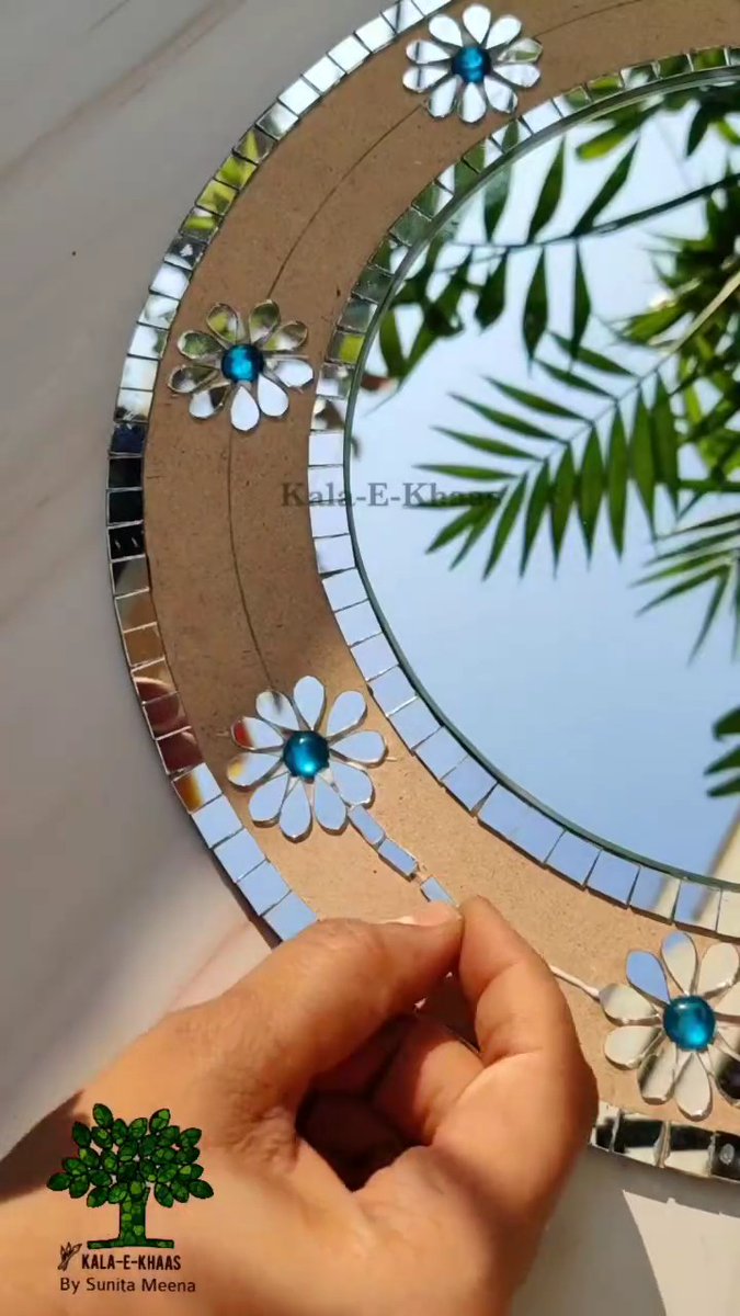 A hand crafting a decorative mirror frame with mirror mosaic tiles and flower designs featuring blue gem centers, branded "KALA-E-KHAAS By Sunita Meena".